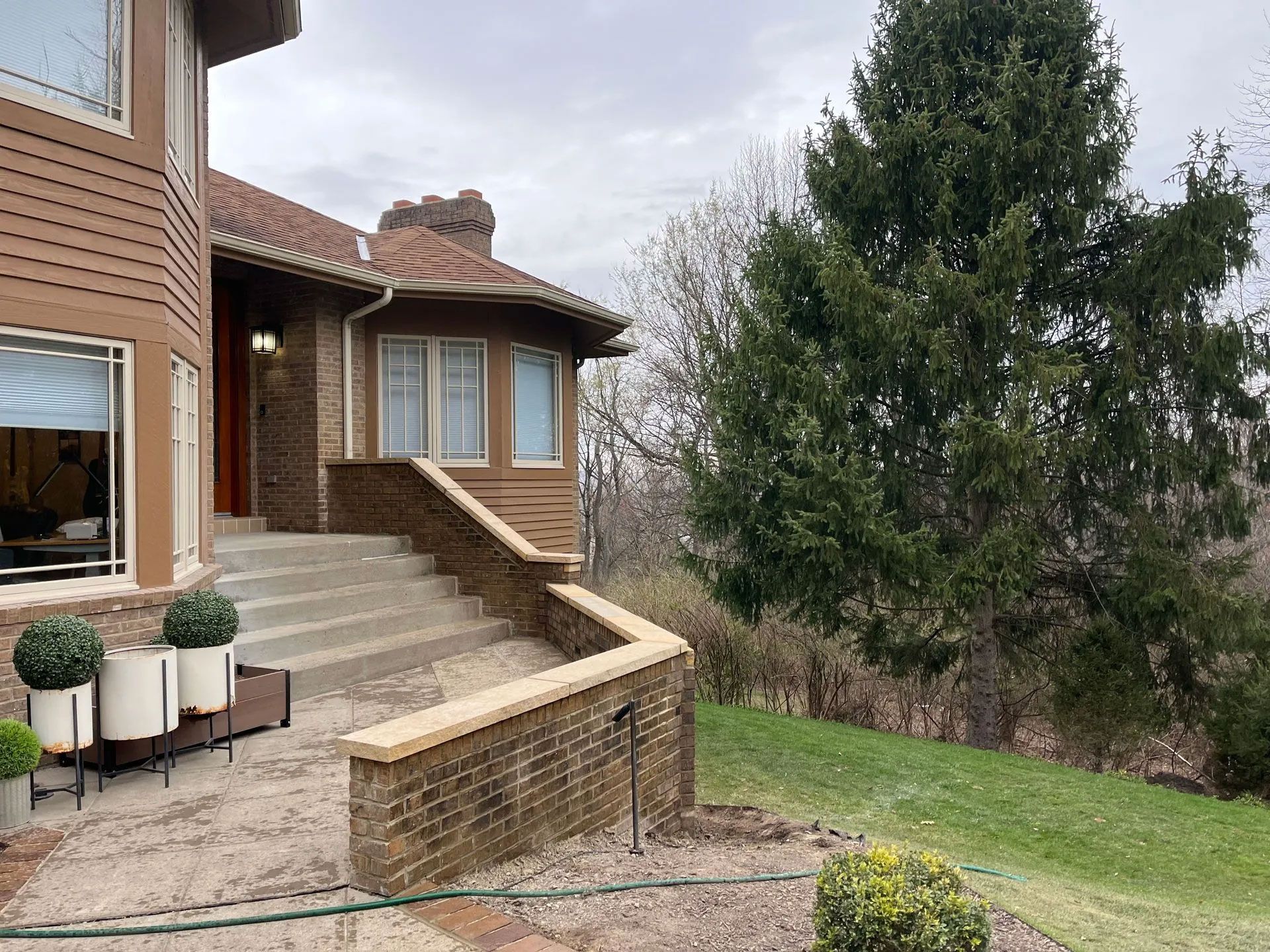Brown house with steps and brick wall. Green lawn and tall evergreen tree. Overcast day.