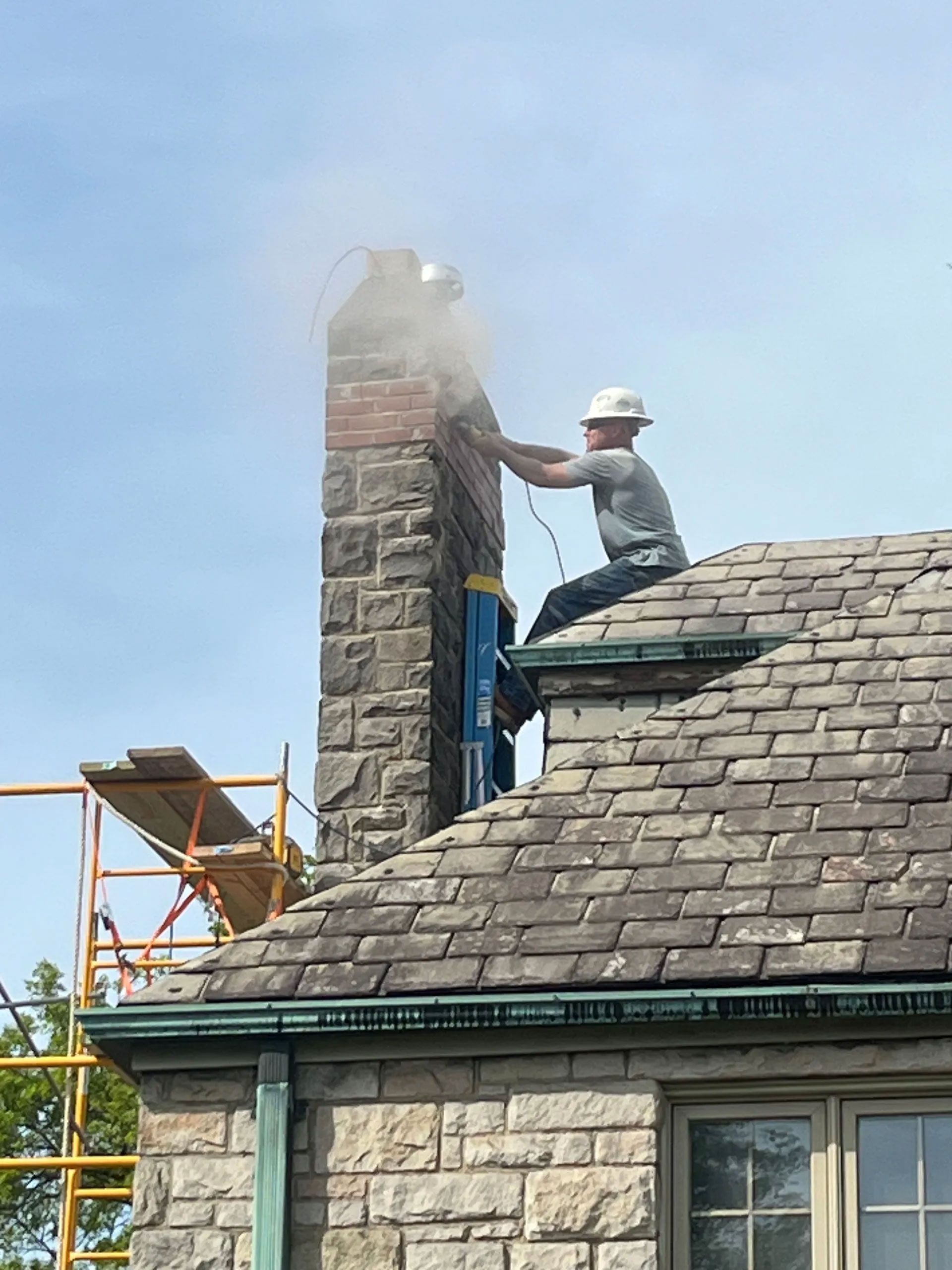 Man on roof using a power saw on a brick chimney, creating dust. Yellow scaffolding, blue sky.