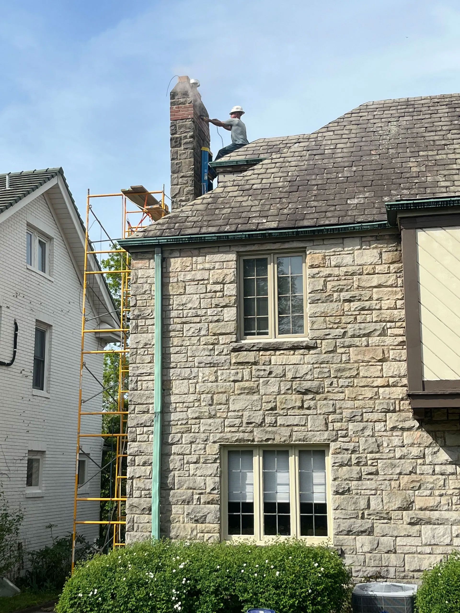 Man on roof cleaning chimney of a stone house with scaffolding, blue sky.