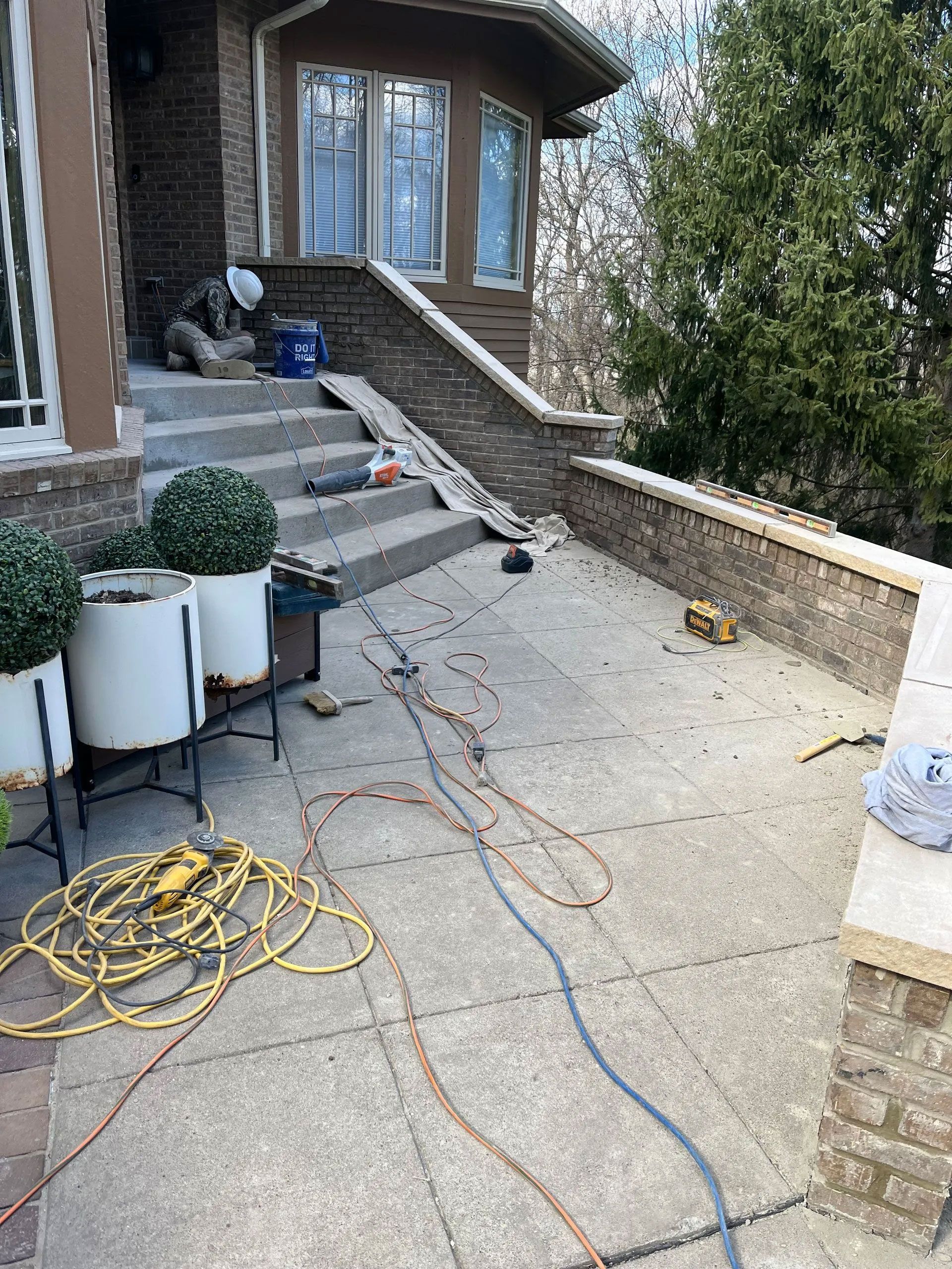 Concrete patio with stone wall and steps, tools and extension cords scattered. Person working in the background.