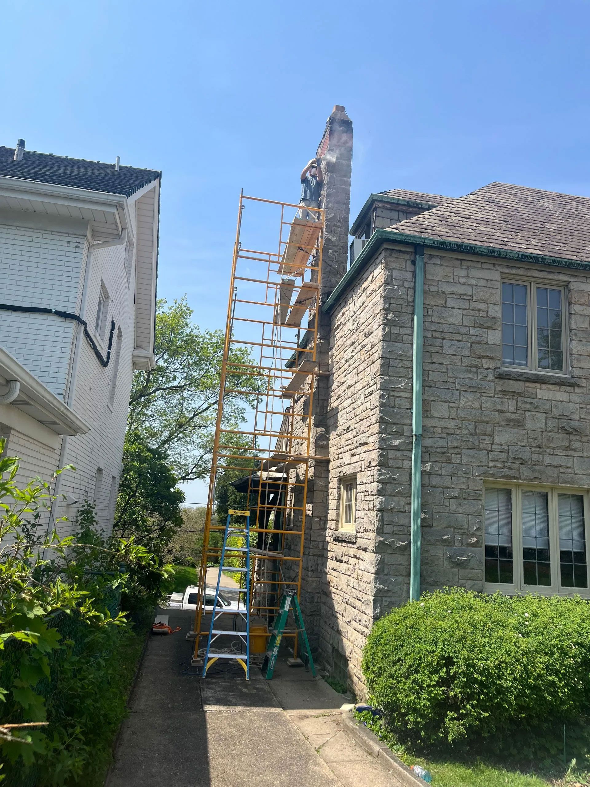 Scaffolding next to a stone building with a chimney and green roof; driveway on the left.