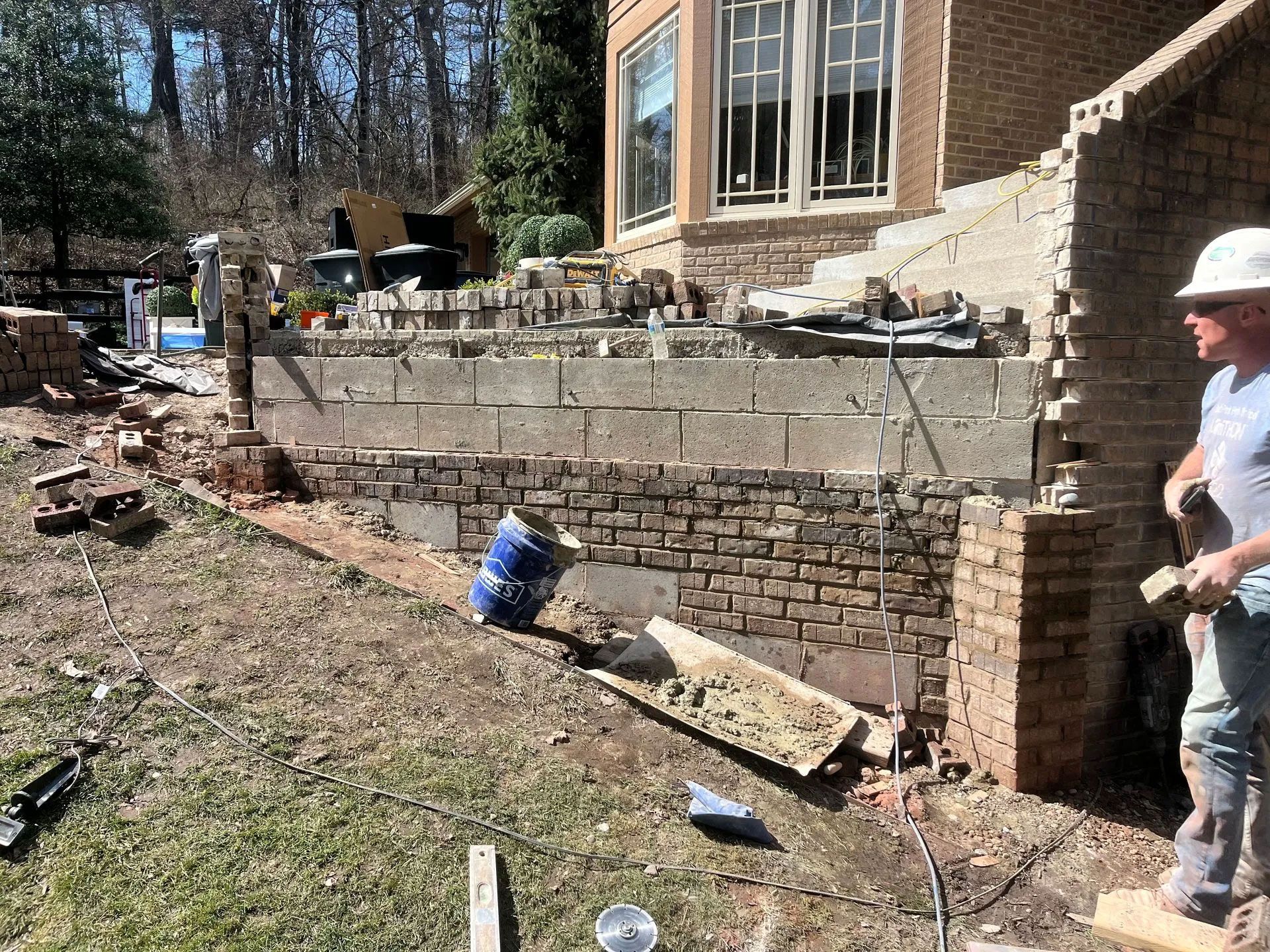 Construction site: partially demolished brick and block wall next to a house; worker in a hard hat.