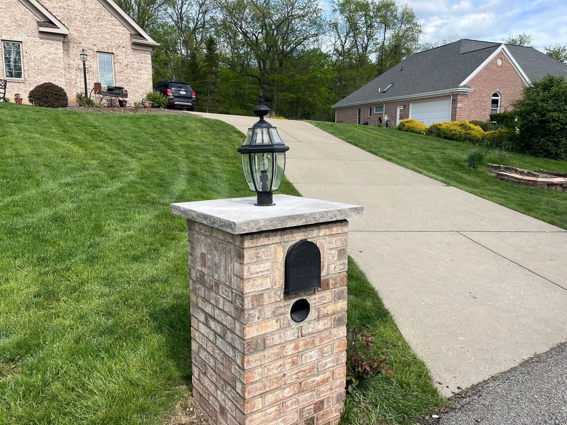 Brick mailbox with lamp on a sloped green lawn, driveway, and two houses in the background.