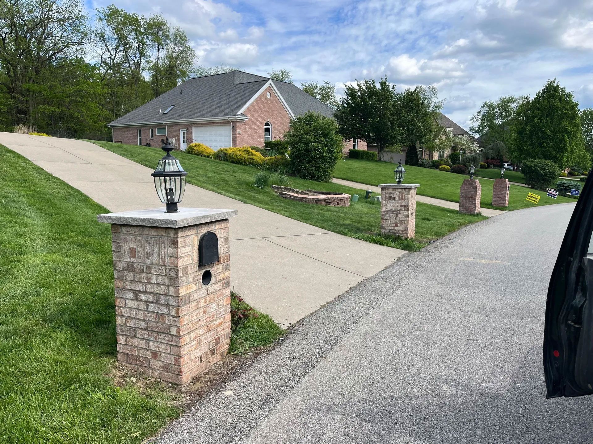 Brick pillars with lamps and mailboxes line a driveway leading to a brick house on a grassy hill.