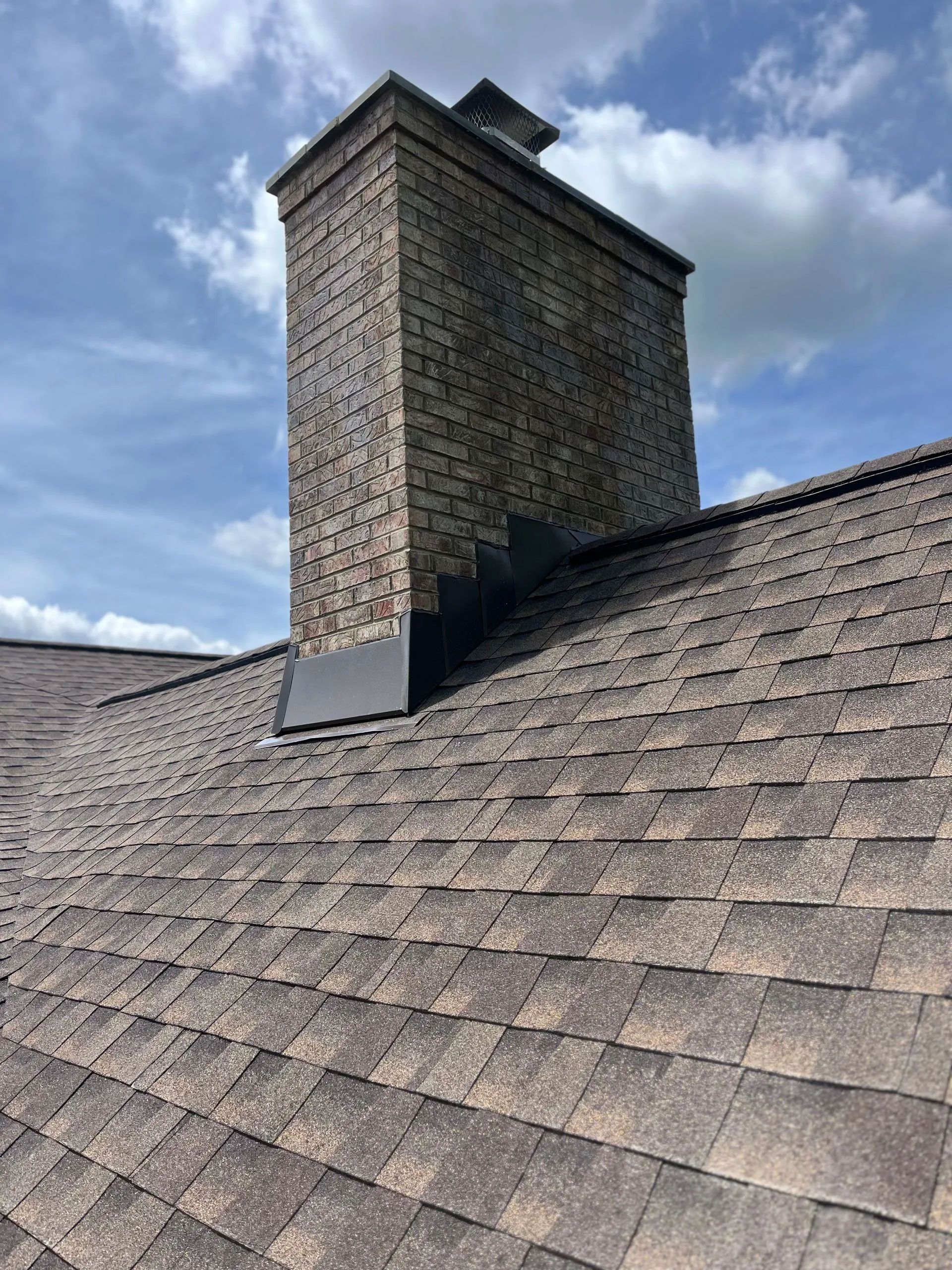 Chimney on a brown shingled roof against a partly cloudy blue sky.
