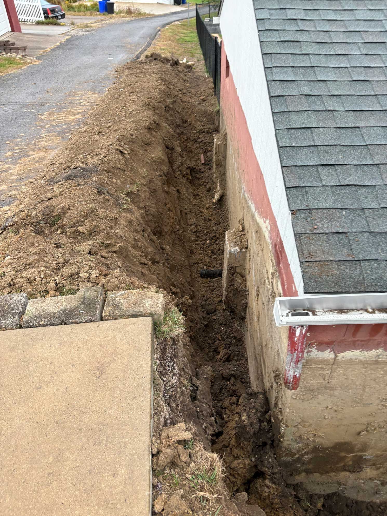 Trench dug next to building. Dirt pile next to concrete and asphalt. Gutter visible.
