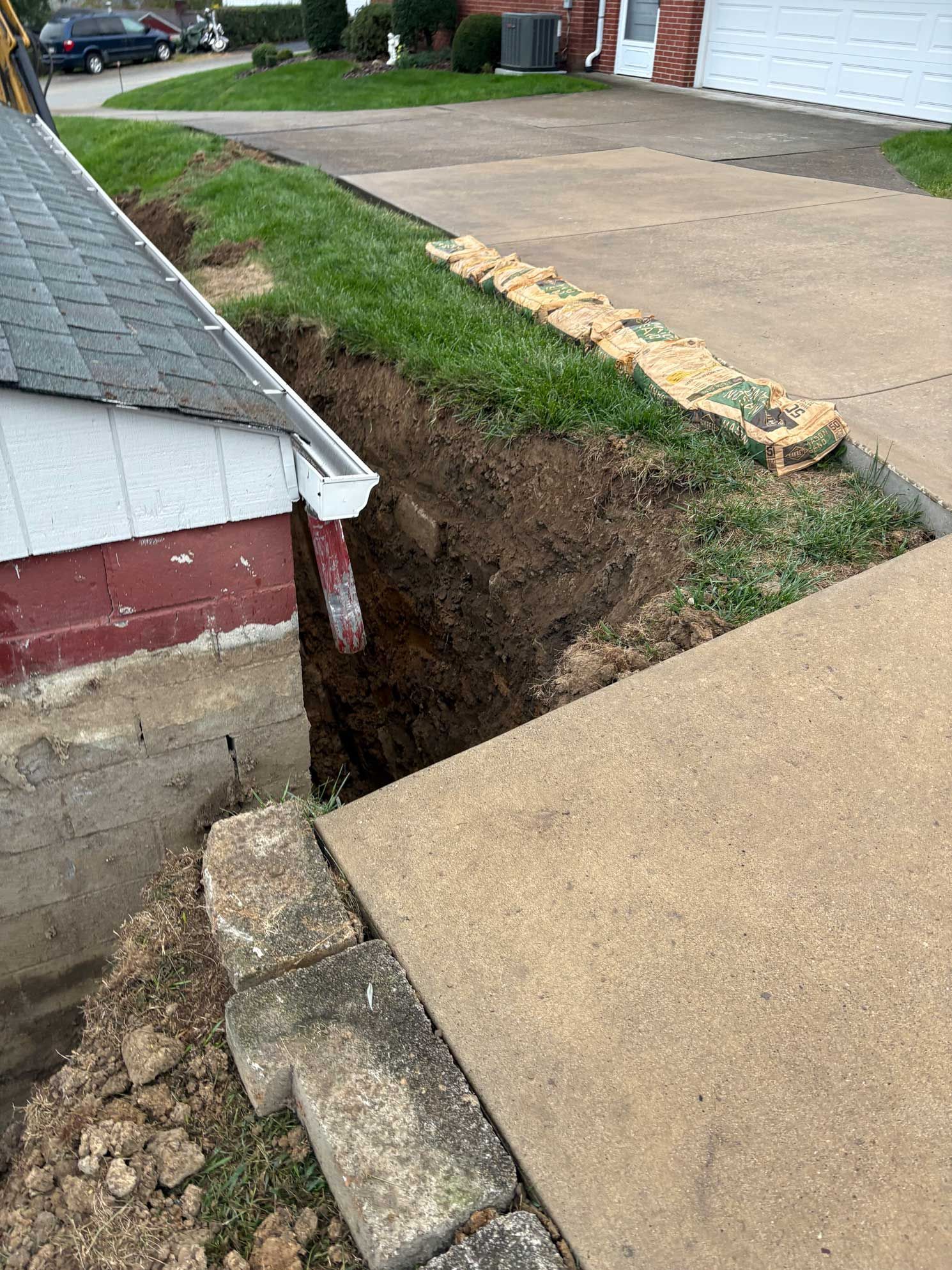 Dug trench next to a garage and sidewalk. Soil alongside a retaining wall. Brown earth visible.