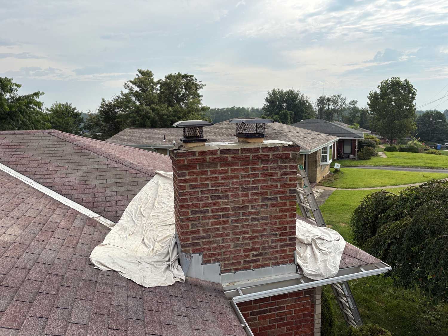 Rooftop view of a brick chimney with two caps. Roofing material is visible; cloudy sky background.