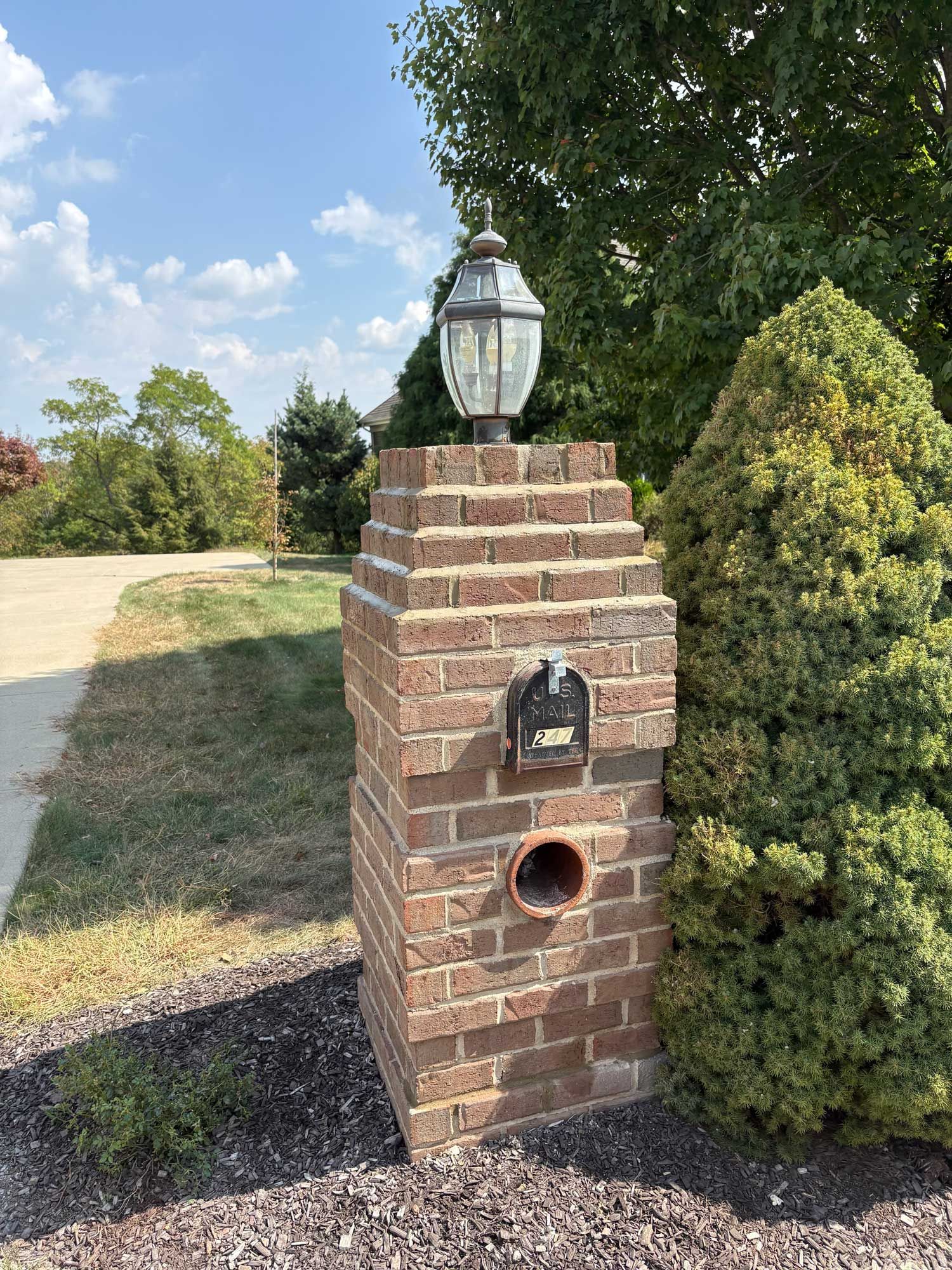 Brick mailbox with lamp, round opening, small address plaque, and green shrubs on a sunny day.