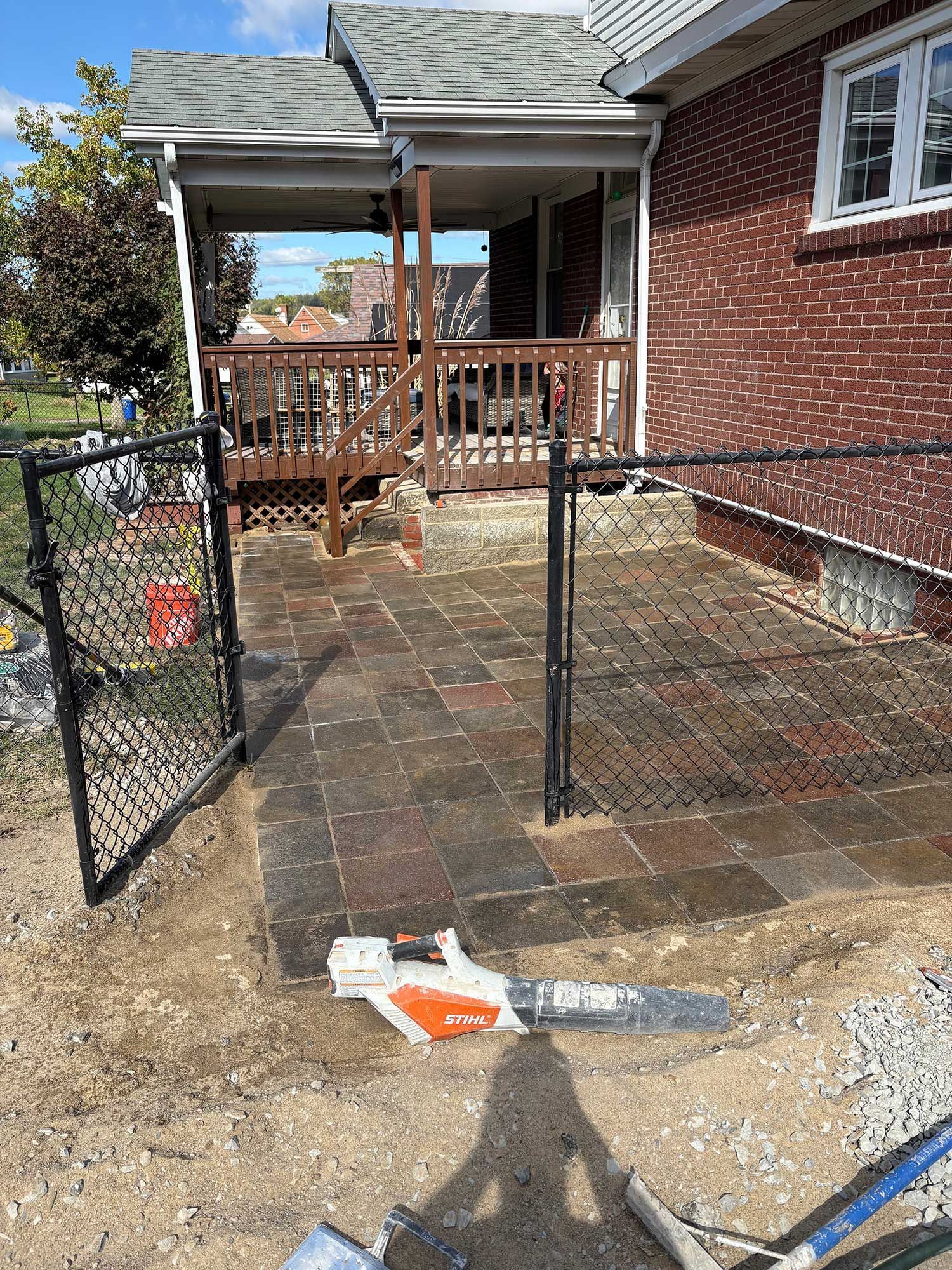 A backyard patio with chain-link fence, brick house, and a chainsaw on the ground.