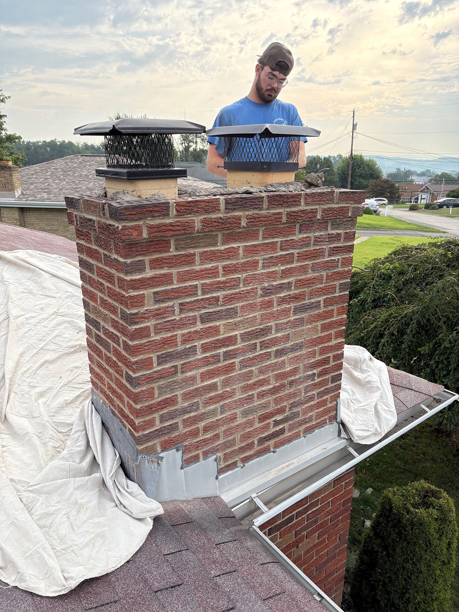 Man on rooftop, installing chimney cap. Brick chimney, tarp covered roof, green background.