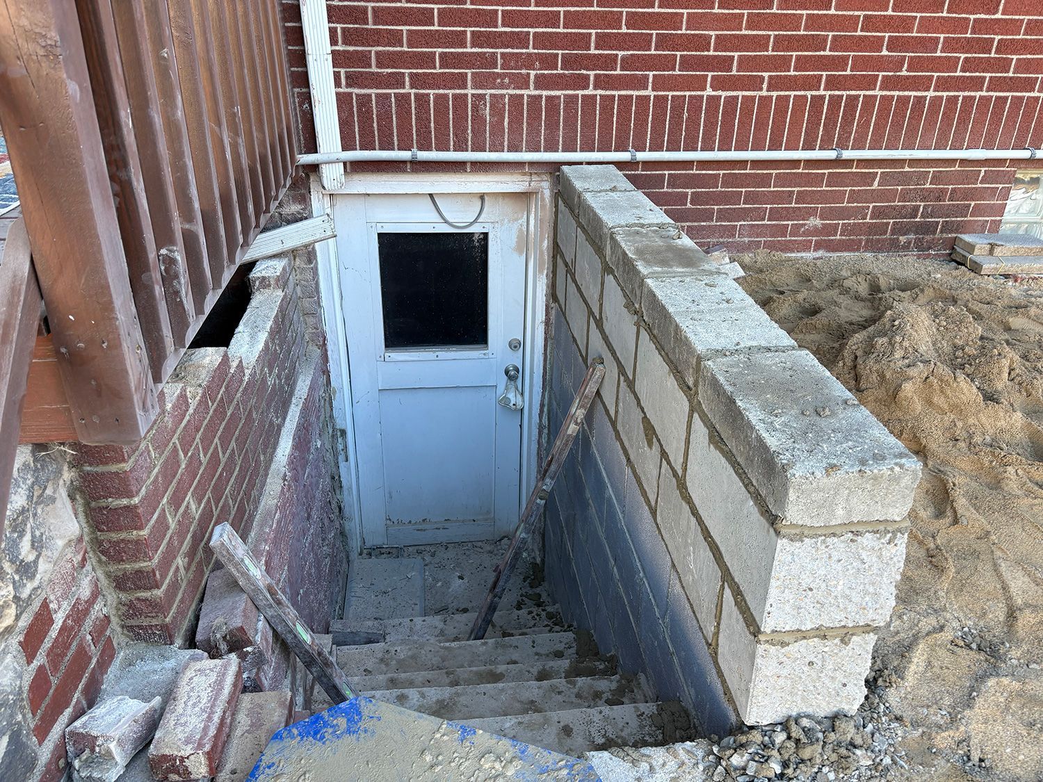 Basement entrance with concrete block retaining wall and steps leading to a white door. Red brick walls, dirt, and construction debris.