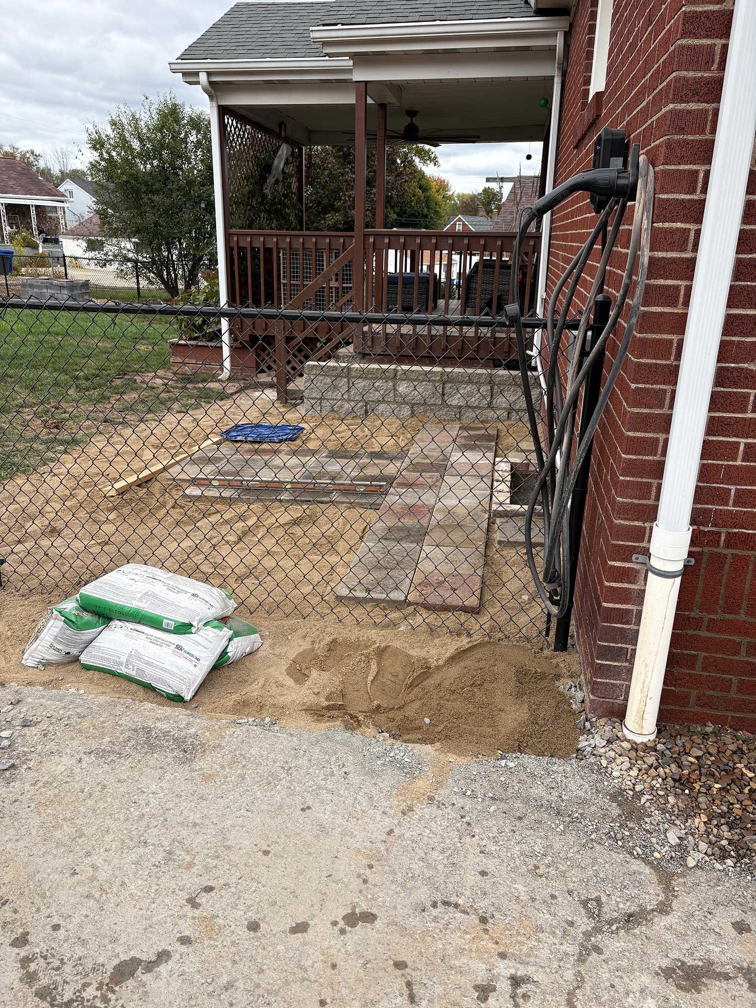 Backyard patio construction: Sand, pavers, fence, porch in background, next to brick building.