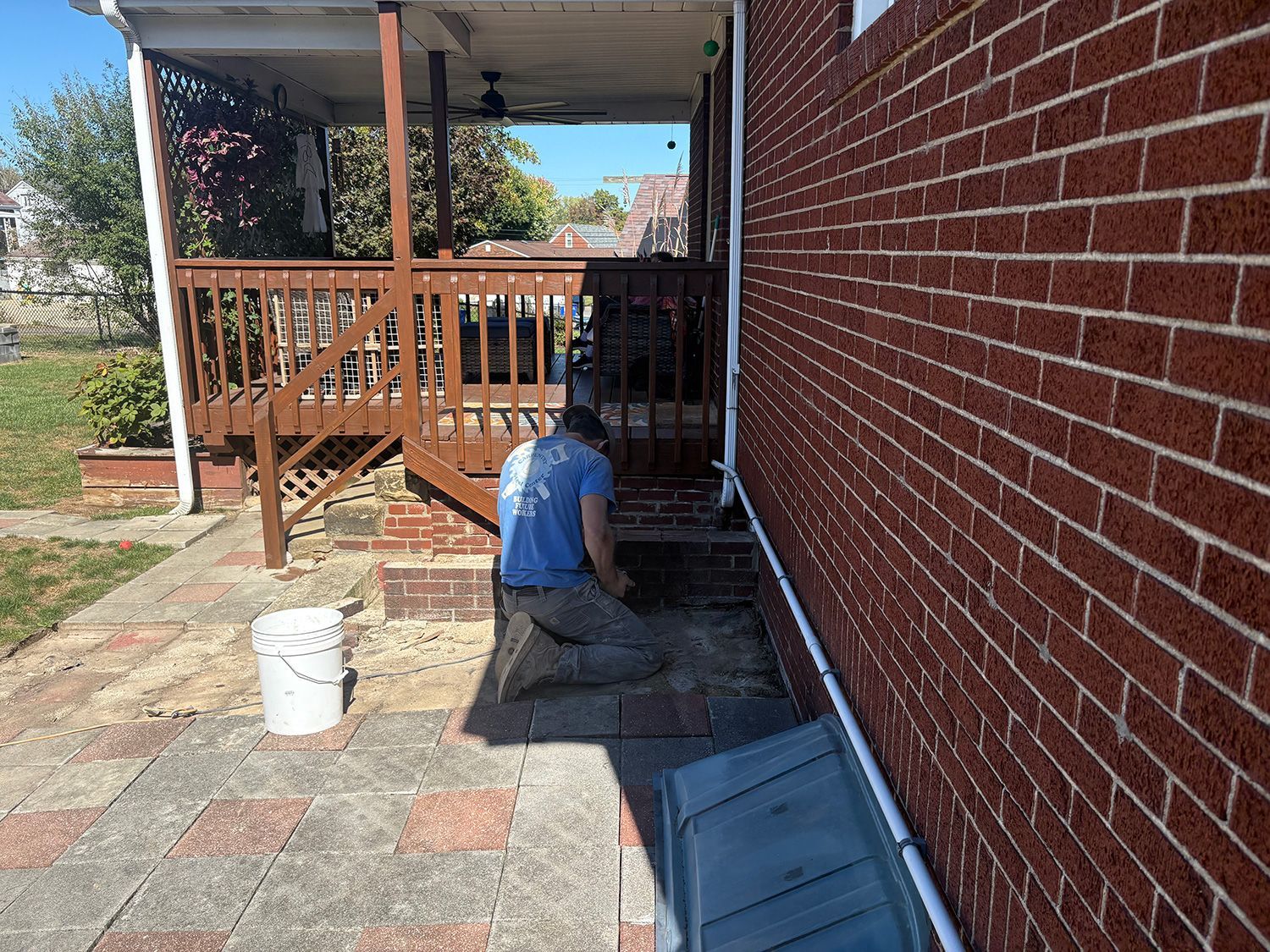 Person kneeling, working near a brick wall and wooden deck. A bucket sits on a stone patio.