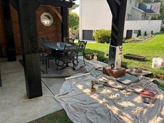 Covered patio with a table, chairs, and tools; tarp and scattered materials on the concrete floor.