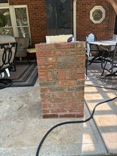Brick patio pillar with a hose draped across a concrete backyard patio near chairs and a house wall