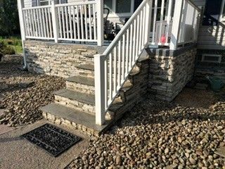 Front porch steps with white railing, stone foundation, and rock landscaping beside a house