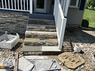 Front steps under construction with stacked stone, white railing, and loose pavers and tools on gravel