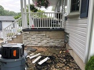 Broken stone wall under a porch railing beside a house, with debris and tools on the ground.