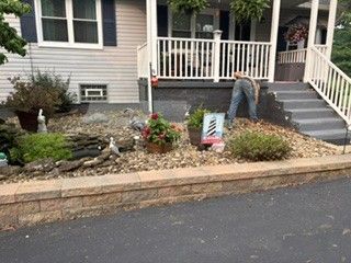 Front yard landscaping with stone retaining wall, shrubs, flowers, and a person bending near the porch steps