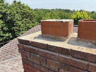 Brick chimney on a rooftop with a forest backdrop. The chimney is topped with a square cap.