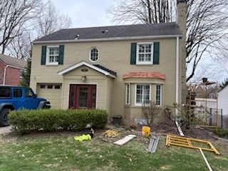 A two-story tan brick house with green shutters, a brown front door, and a bay window, with equipment on the front lawn.