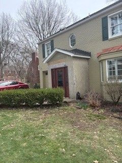 A two-story tan brick house with a front porch, dark shutters, and a well-manicured hedge in the front yard.