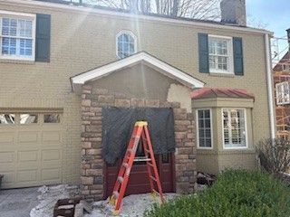 A red ladder stands before a house entrance under construction with stone veneer partially installed on the porch.