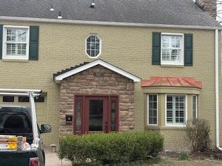 A two-story tan brick house with a stone-accented front entryway, dark shutters, and a bay window under a copper roof.