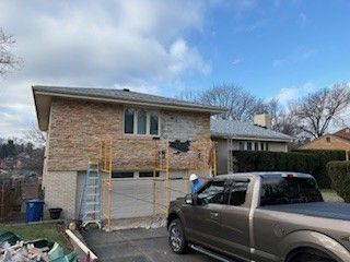 A worker in a hard hat stands by a brown truck at a home with a partial stone facade being repaired on scaffolding.