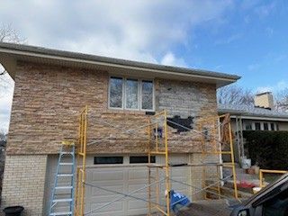Exterior of a two-story home with partial tan stone siding, a garage, and yellow scaffolding for exterior repairs.