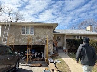 Workers on scaffolding repair the exterior stucco wall of a suburban house on a sunny day.