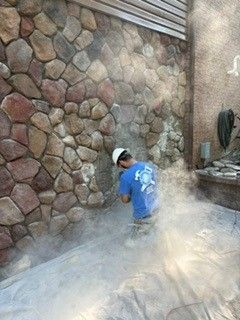 Worker power-washing a stone wall, with water spraying across the ground.