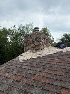 Rooftop with a stone chimney and loose shingles under a cloudy sky, surrounded by trees
