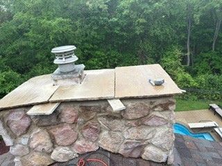 Stone chimney with metal vent and tan cap beside a pool, with dense green trees in the background
