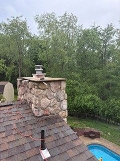 Backyard chimney and shingled roof beside a blue pool, with trees in the background