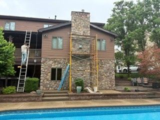 Two workers on ladders repairing a stone house beside a swimming pool