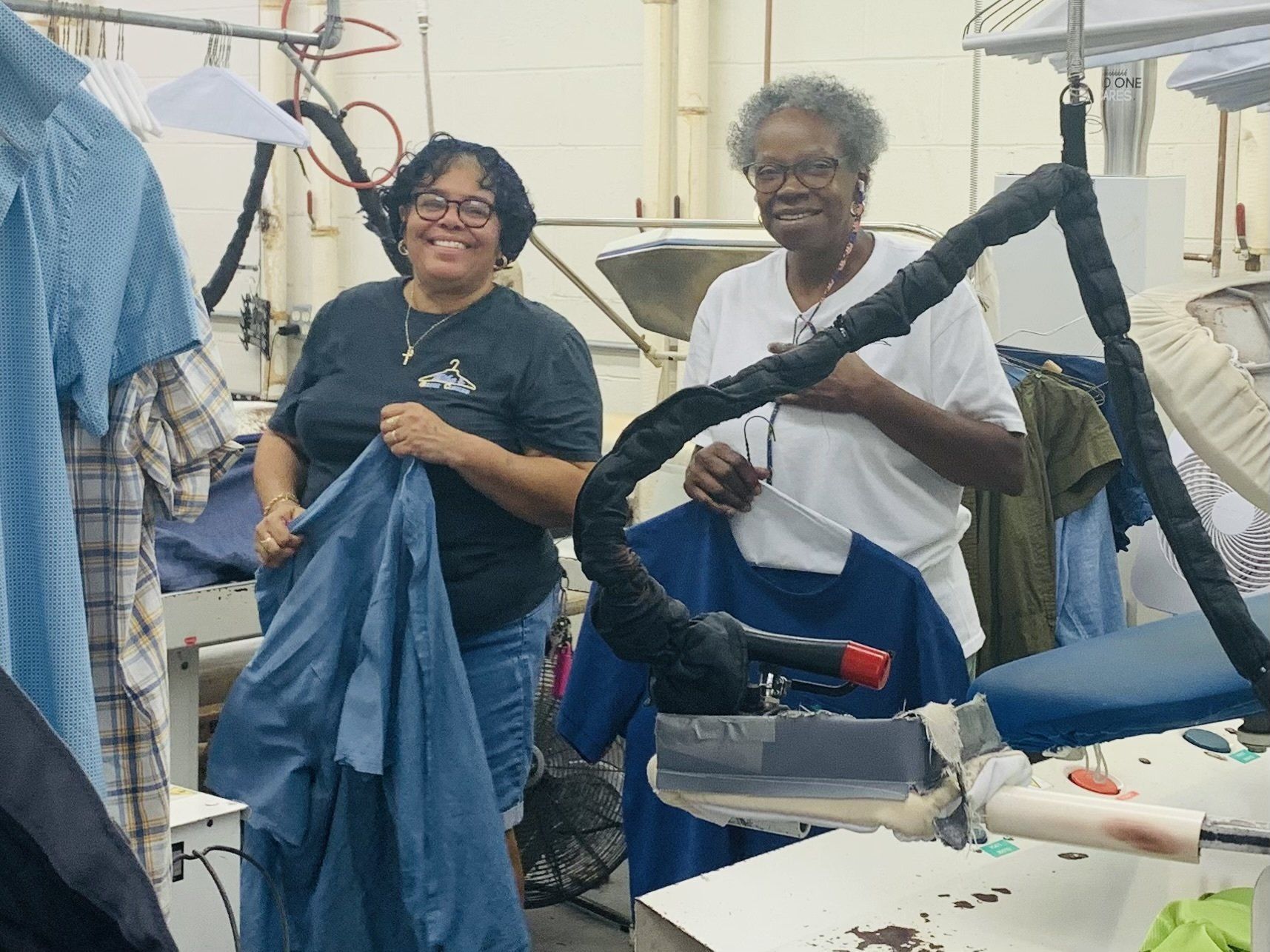 Two people smiling while folding clothes in a laundry room, with hanging garments and laundry equipment nearby