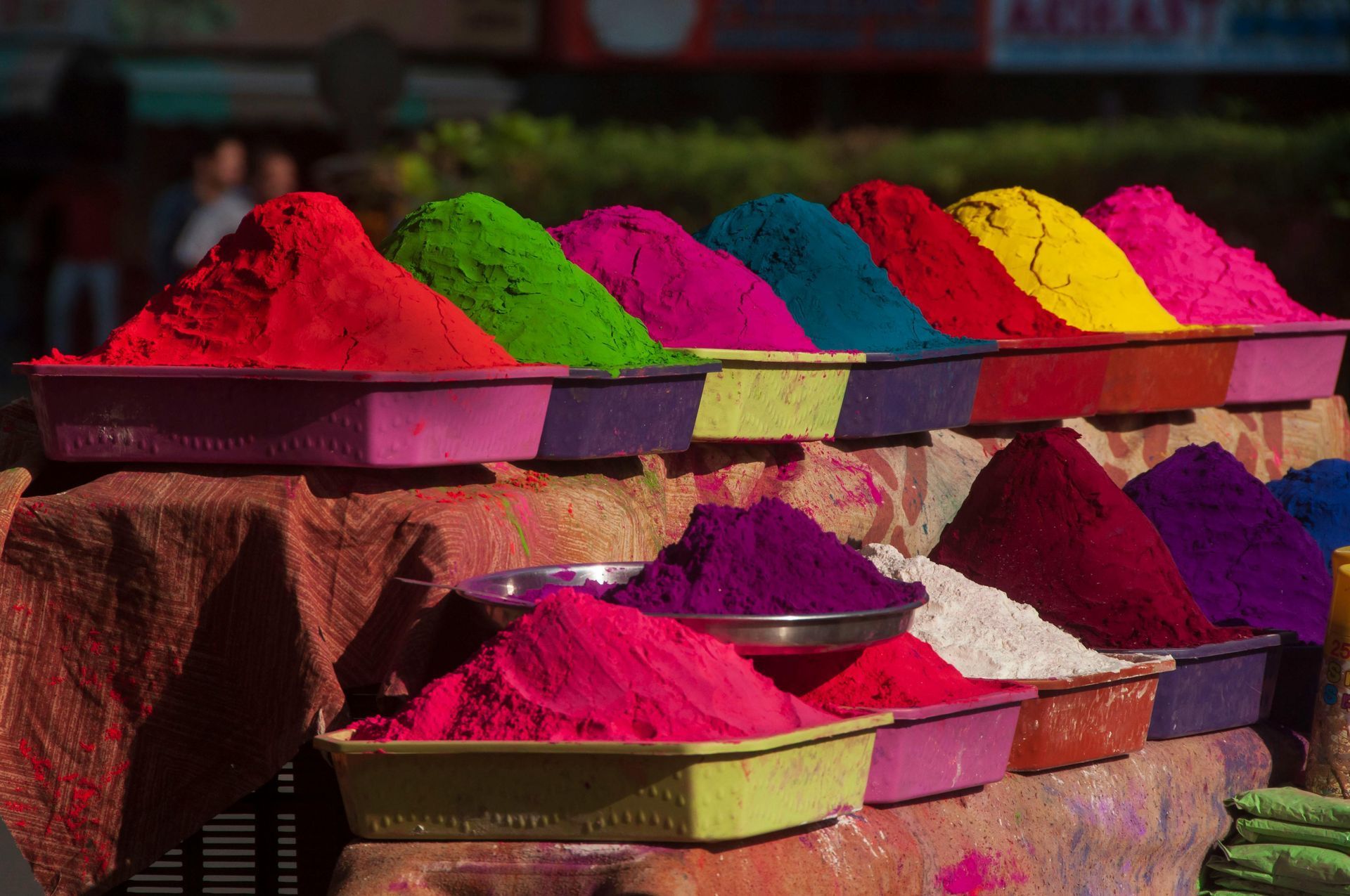 Bright piles of colorful powders at a market stall, including red, green, yellow, blue, and pink.