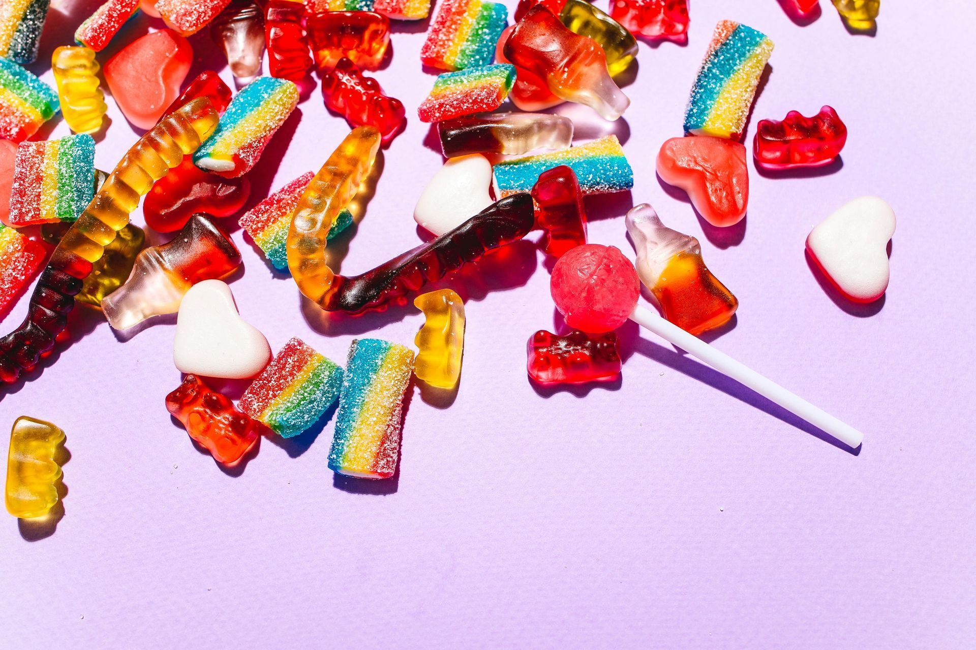 Colorful gummy candies scattered on a lavender surface with a white lollipop stick