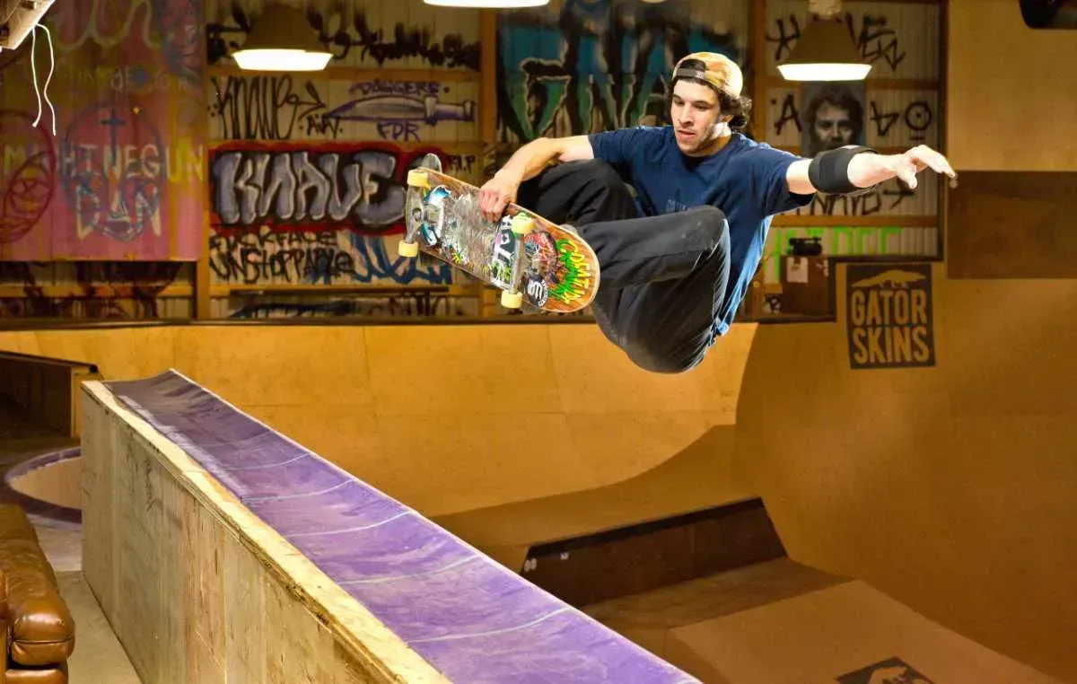 Skateboarder airborne over a ramp in an indoor skate park, board angled up amid graffiti-covered walls.
