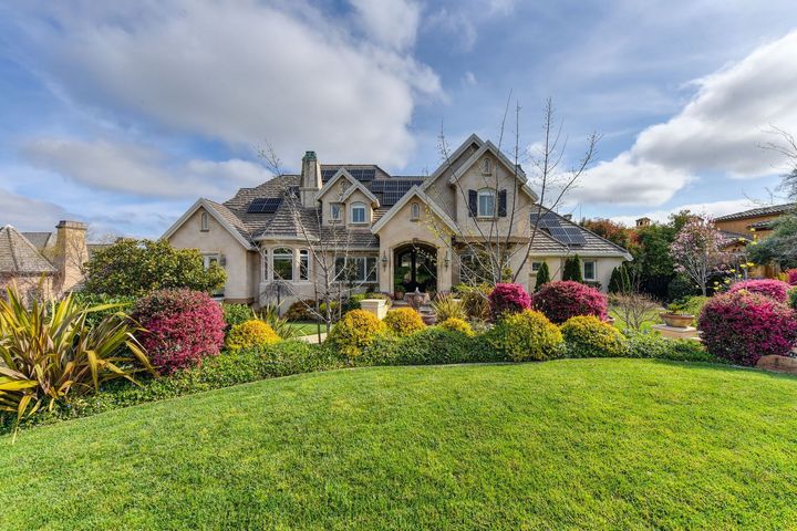 Stone house with gabled roofs and a lush flower garden under a partly cloudy sky