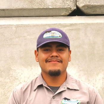 Smiling person in a purple cap and gray shirt standing against a concrete wall