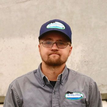 Man wearing a blue cap and gray button-up shirt, standing against a plain wall.