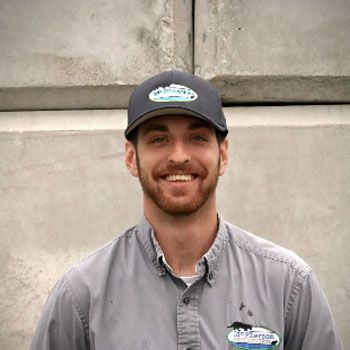 Smiling man in a gray shirt and dark cap standing against a concrete wall