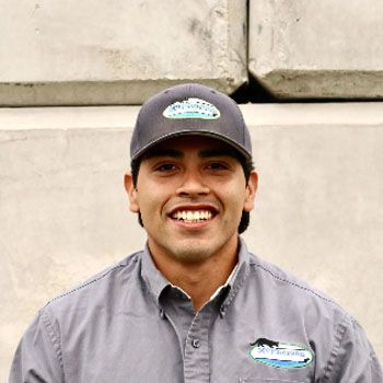Smiling person in a gray shirt and cap standing against a light concrete wall.