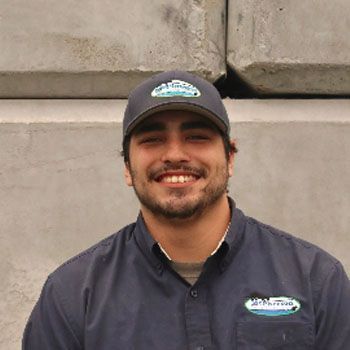 Smiling man in a dark cap and shirt standing against a concrete wall.