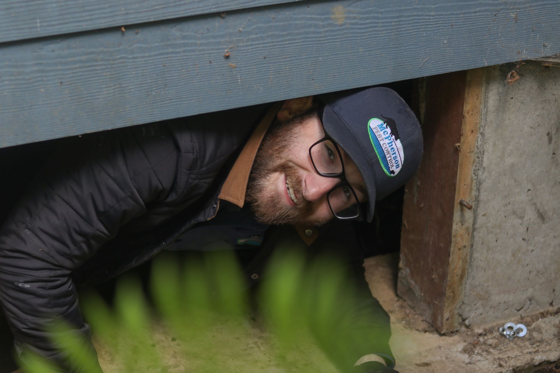 Person in a cap and glasses peeks through a low crawlspace opening under a building.