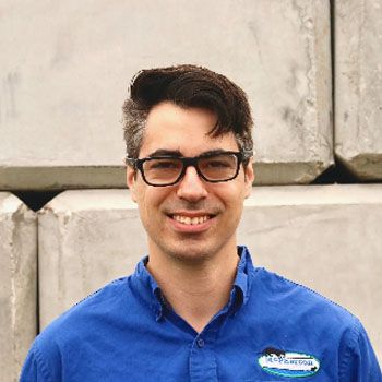 Smiling person with black glasses and a blue collared shirt standing against a concrete wall