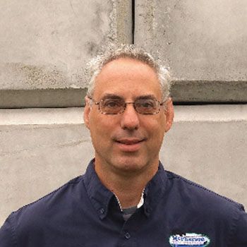 Man standing against a concrete wall, wearing glasses, a navy shirt, and a slight smile.